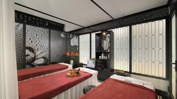 A spa room with two red-towel-covered beds, a decorative wall with water droplets, and a window with a geometric pattern.