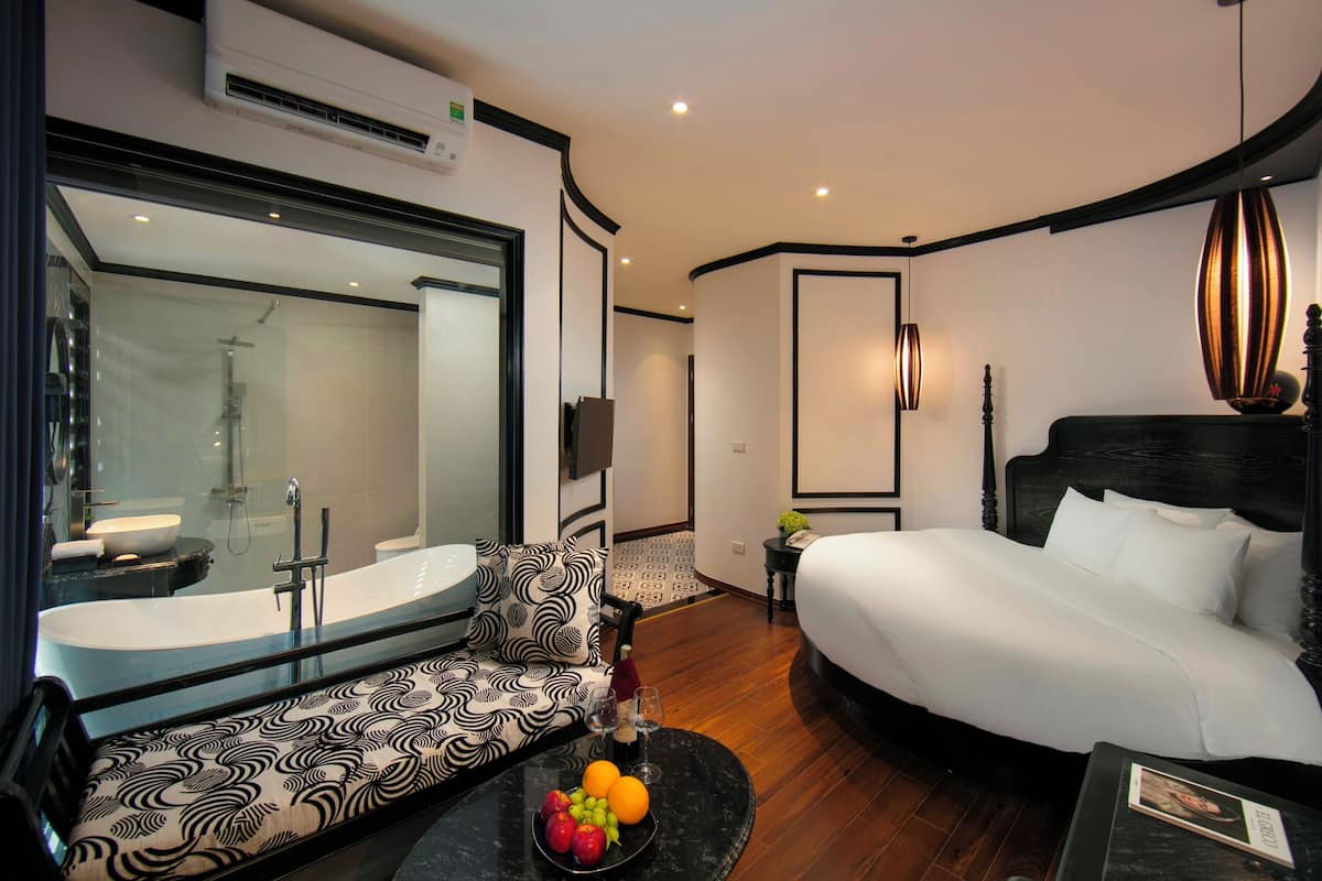 Una habitación de hotel moderna con una cama grande, un sofá, una mesa redonda con fruta y un baño con bañera y ducha.