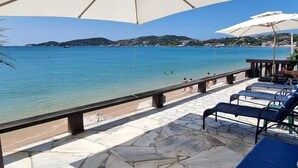 Terrassa de pedra amb seients de saló i ombrel·les, amb vista a una platja d'aigua turquesa i turons allunyats.