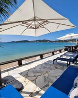 Un patio con suelo de piedra, sillones y sombrillas, con vista a una playa de aguas azules cristalinas y colinas lejanas.