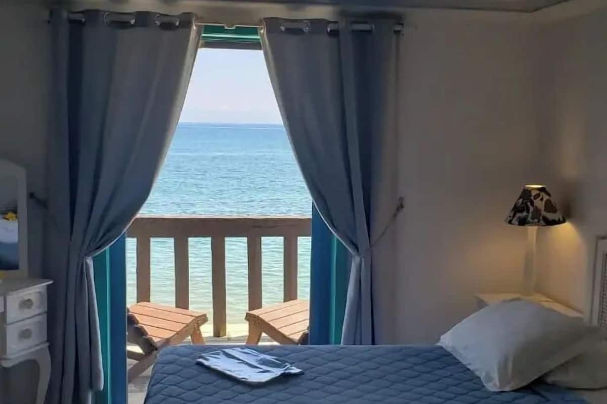 A bedroom with a bed, a nightstand, a lamp, and a view of the sea through curtains.