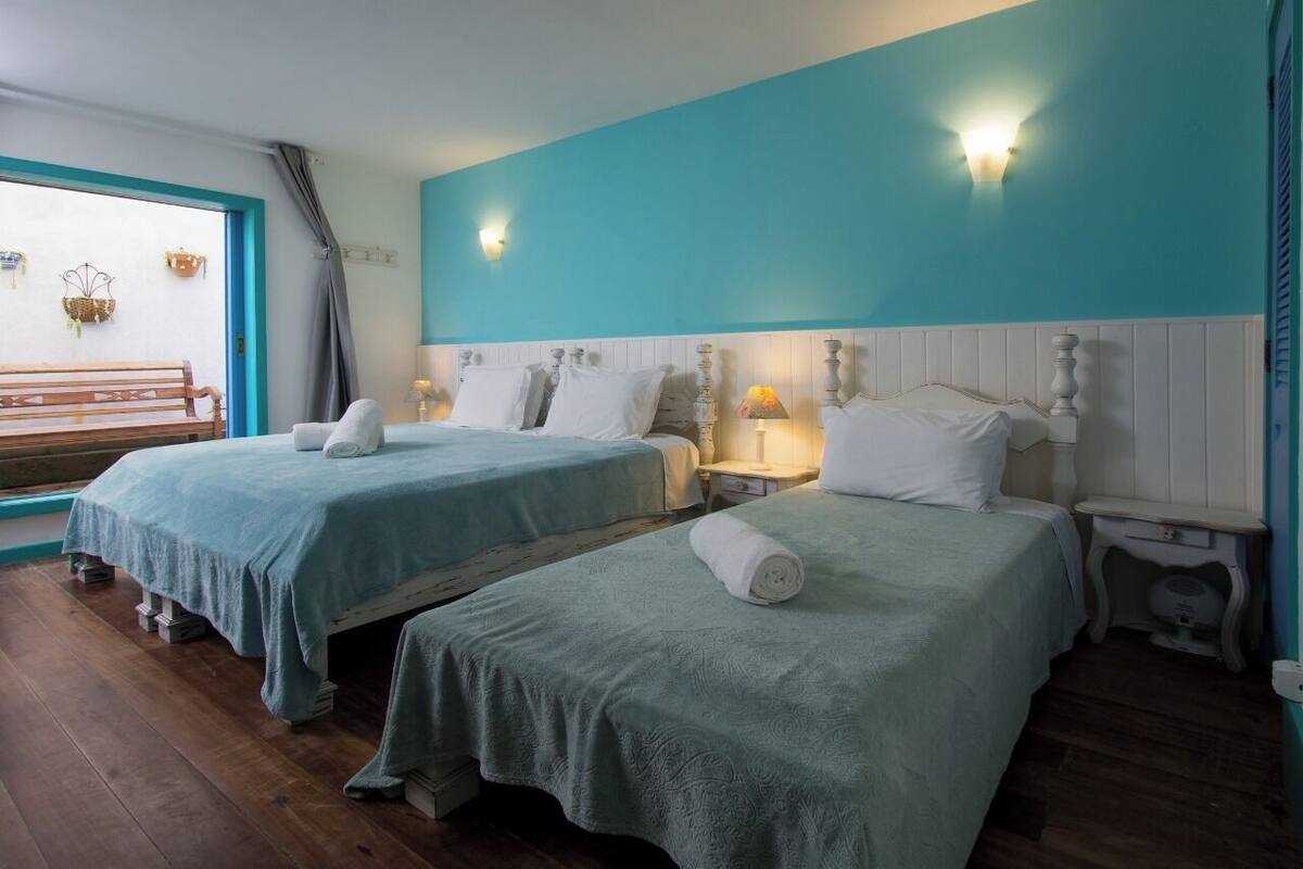 A room with two beds, each with a white headboard and a teal wall. There are bedside tables with lamps and a bench in the corner.