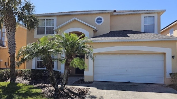 A two-story house with a white garage door, a small front yard with palm trees, and a clear blue sky. - Spacious 7 Br, 4.5 Br Retreat With Pool 7 Bedroom Condo by RedAwning (Kissimmee)