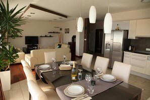 Un espace repas moderne avec une table en bois foncé, dressée pour quatre personnes, entourée de chaises à revêtement blanc. Une grande plante en pot est visible sur la gauche, et un coin cuisine avec des appareils en acier inoxydable se trouve à l’arrière-plan.