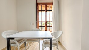 Una habitación con una mesa blanca y cuatro sillas de mimbre, una puerta de madera con paneles de vidrio y vistas a zonas verdes al exterior.
