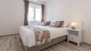 A bedroom with a bed, a nightstand with a lamp, a window with curtains, and a wooden floor. - Apartments Oleander Sea View (Konavle)