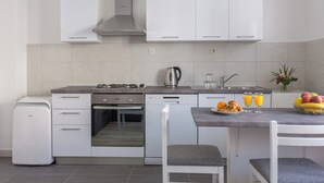 A modern kitchen with white cabinets, a built-in oven, a dishwasher, and a coffee maker. There is a dining table with chairs and a bowl of fruit. - Apartments Oleander Sea View (Konavle)