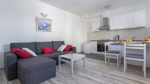 A modern living room with a grey sofa, a small white table, and a kitchen area with white cabinets and a dining table. - Apartments Oleander Sea View (Konavle)