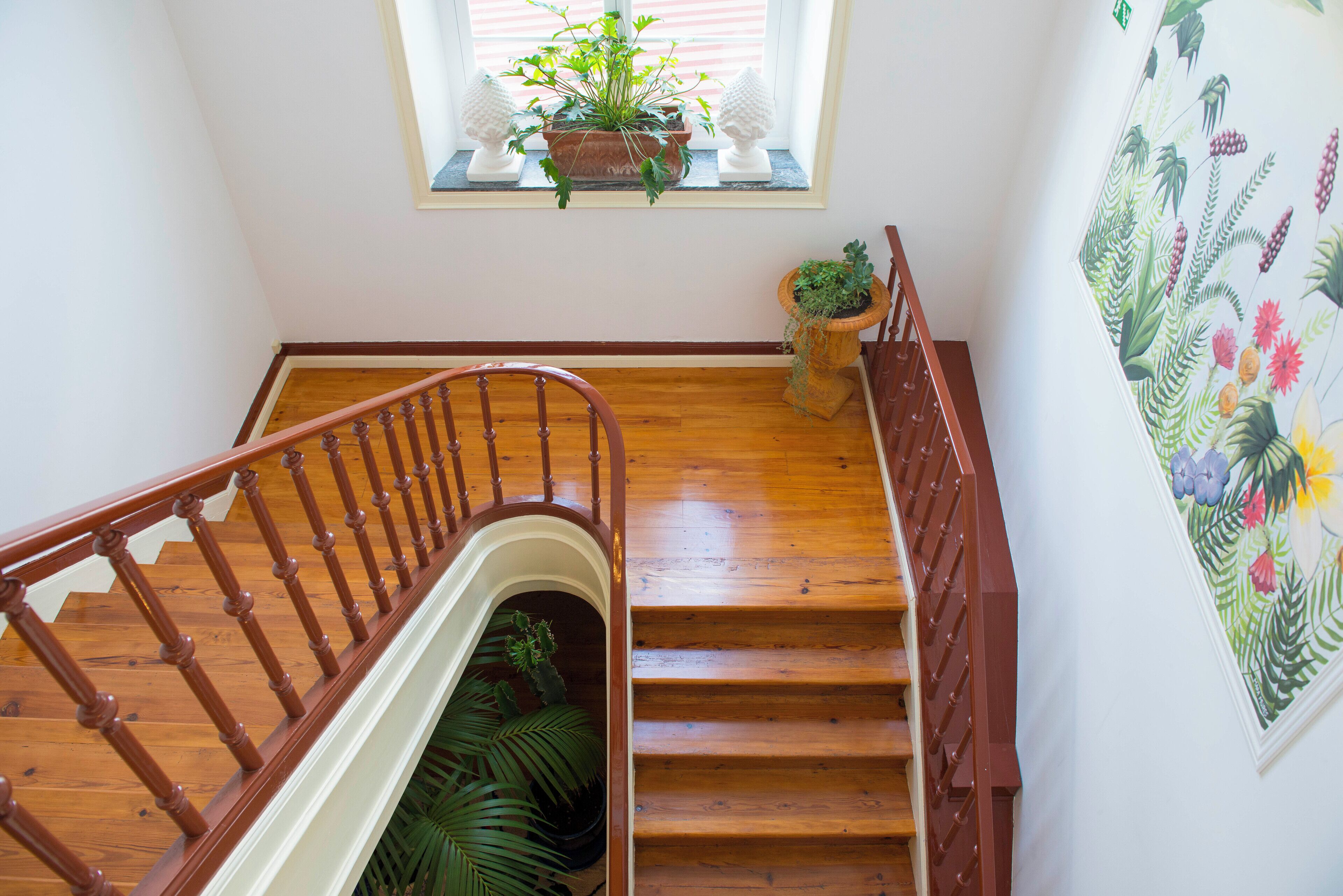 Una escalera de madera con barandilla, una ventana con planta y una pared decorada con un mural floral.