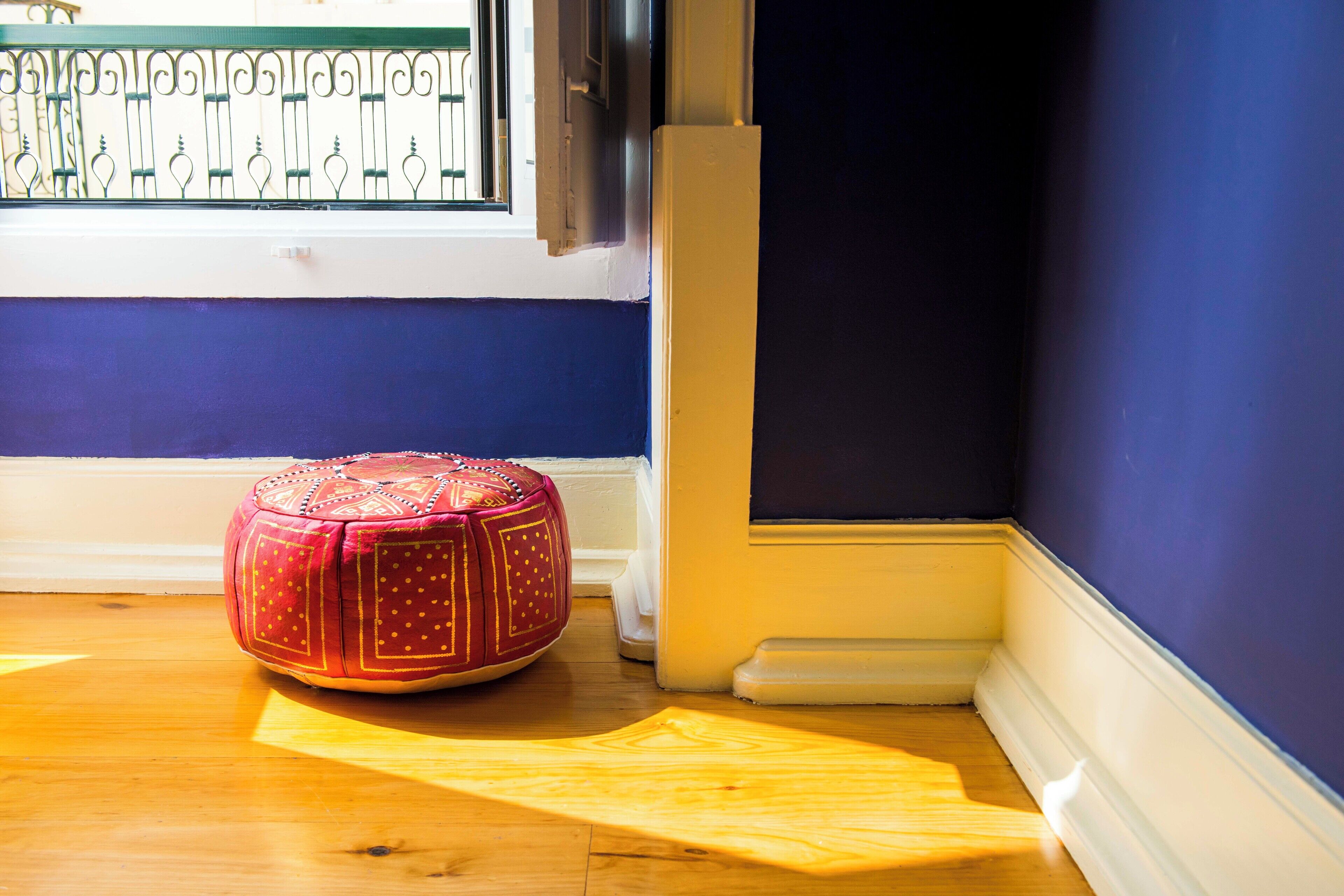 A room with a wooden floor, a red ottoman, a window with metal bars, and dark blue walls.