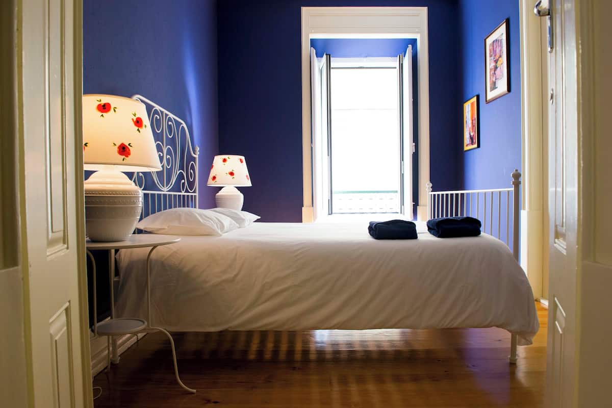 A bedroom with a bed, two bedside lamps, a white table with a bowl, and a door leading to a balcony.