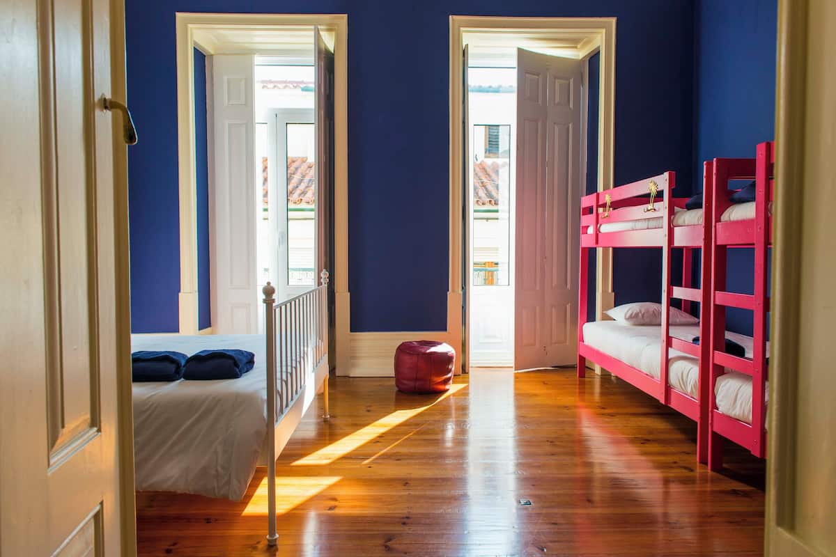 A room with a bunk bed, a single bed, and a red ottoman. There are two doors leading to balconies.