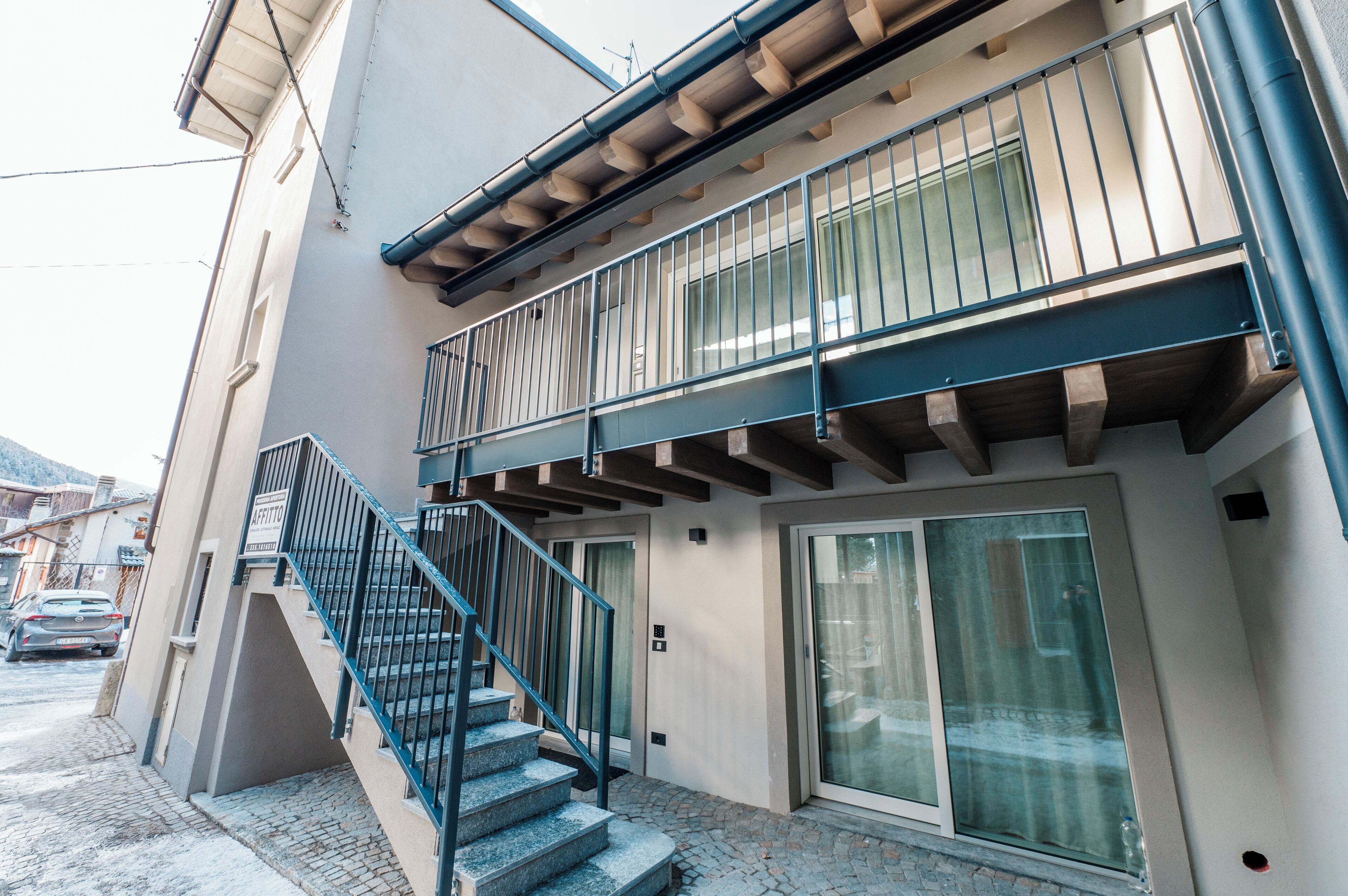 A two-story building with a staircase leading to the entrance, a balcony with metal railings, and a sliding glass door.