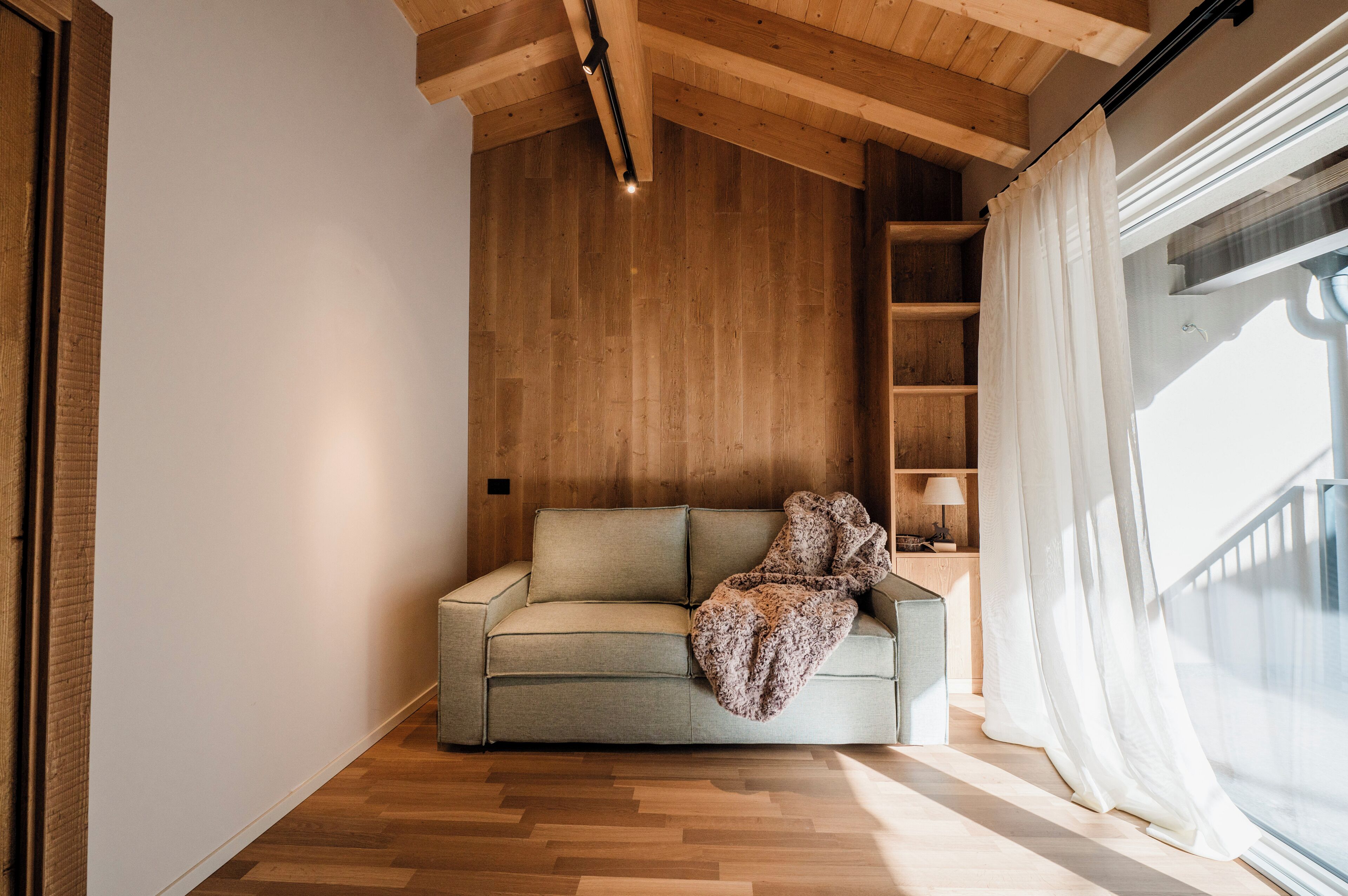 Um quarto aconchegante com teto de madeira, um sofá com manta, uma estante de madeira e uma janela com cortinas.