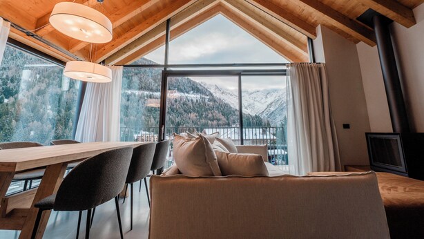 A cozy room with a wooden ceiling, large windows, a dining table, and a sofa. - DALANIA MOUNTAIN APARTMENTS (Ponte di Legno)