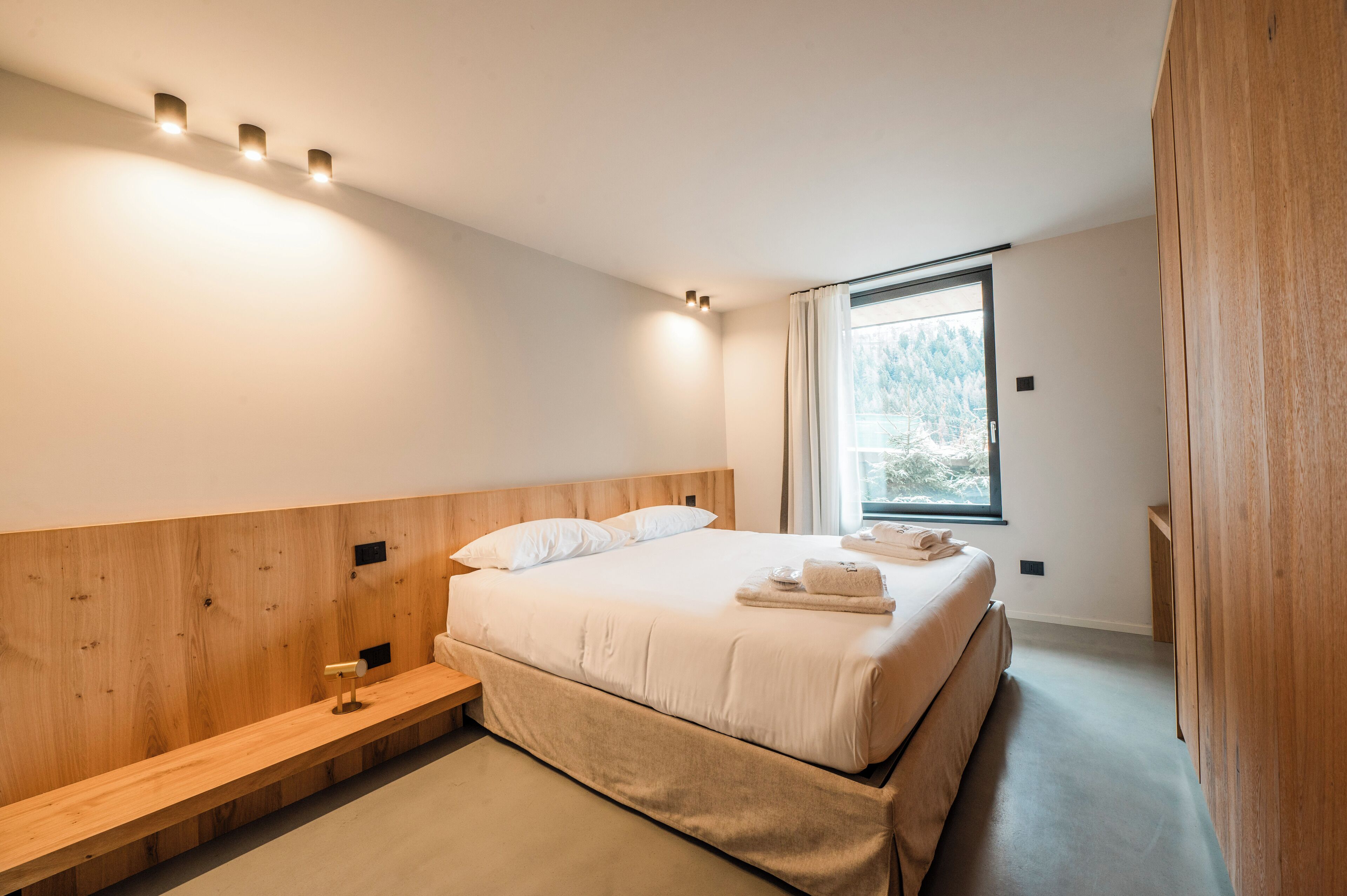 Ein modernes Schlafzimmer mit einem großen Bett, einem Holz-Kopfteil, Nachttischen und einem Fenster mit Blick auf Bäume.