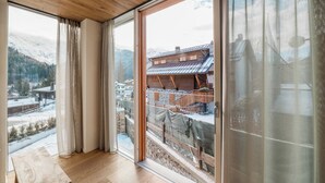 A room with wooden flooring, large windows with curtains, and a view of a snowy mountain and traditional buildings. - DALANIA MOUNTAIN APARTMENTS (Ponte di Legno)