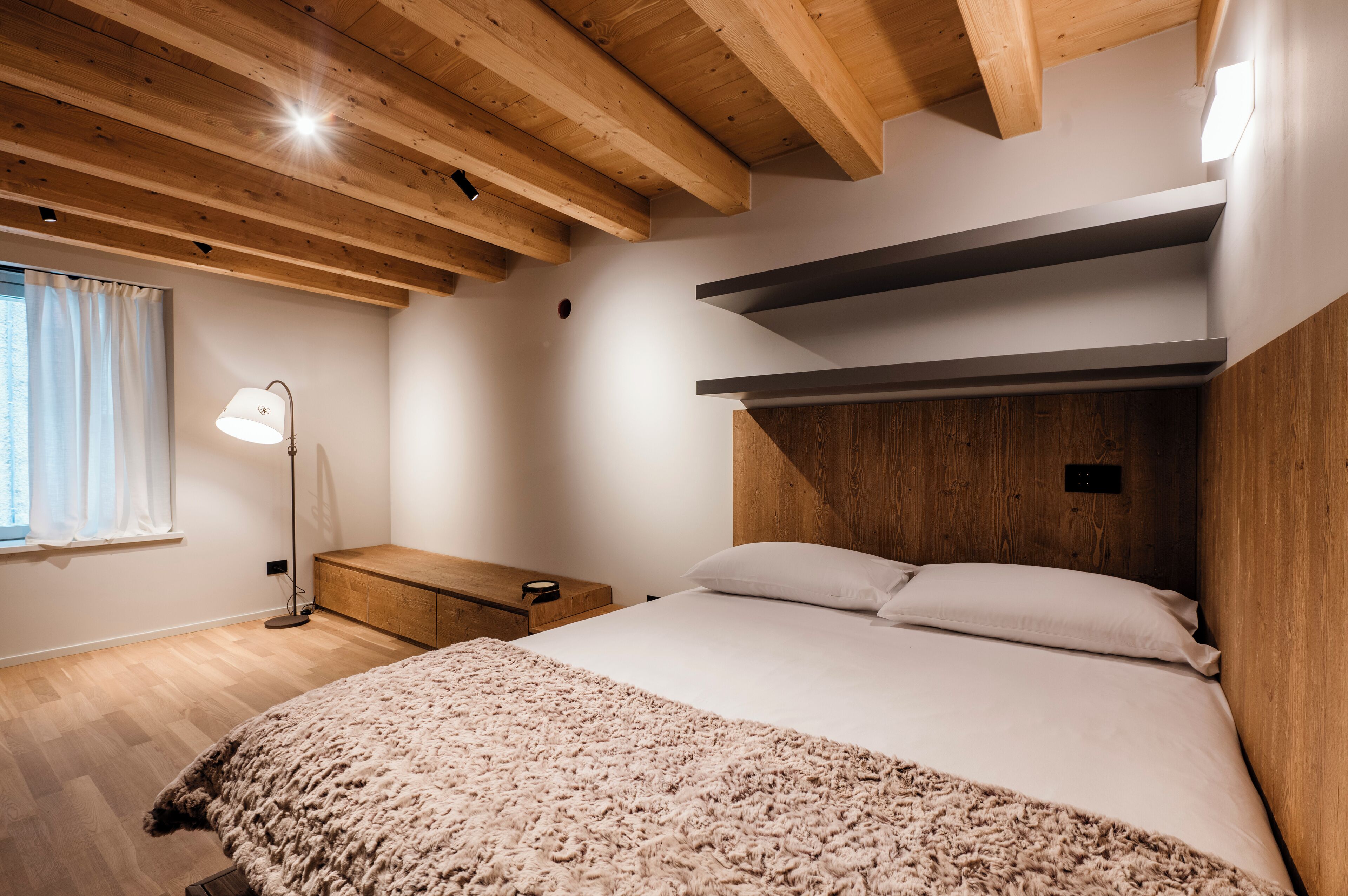 A bedroom with a wooden ceiling, a bed with white bedding, a nightstand, and a floor lamp.