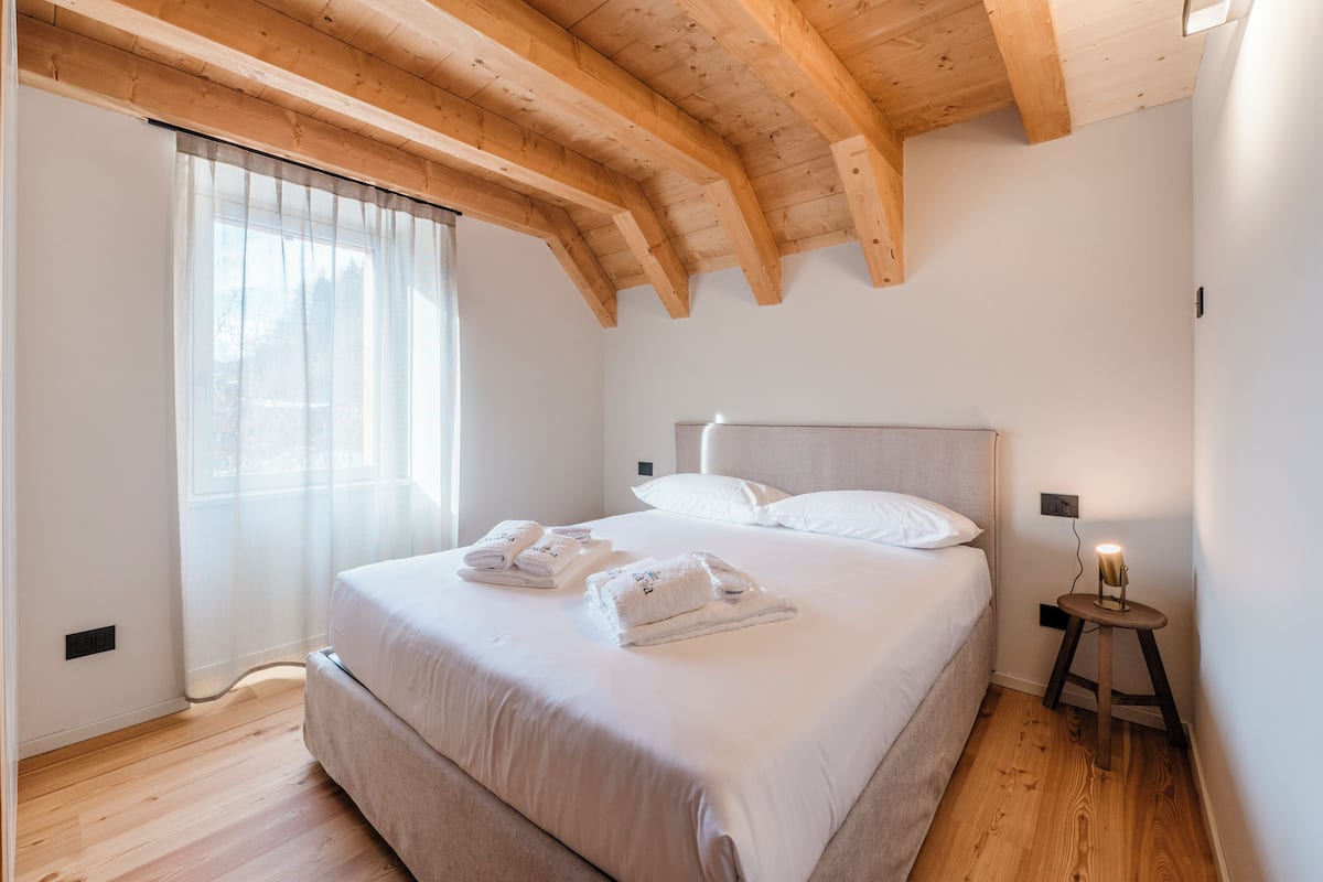 Un dormitorio con techo de madera, una cama con sábanas blancas, una mesita de noche con lámpara y una ventana con cortinas de encaje.