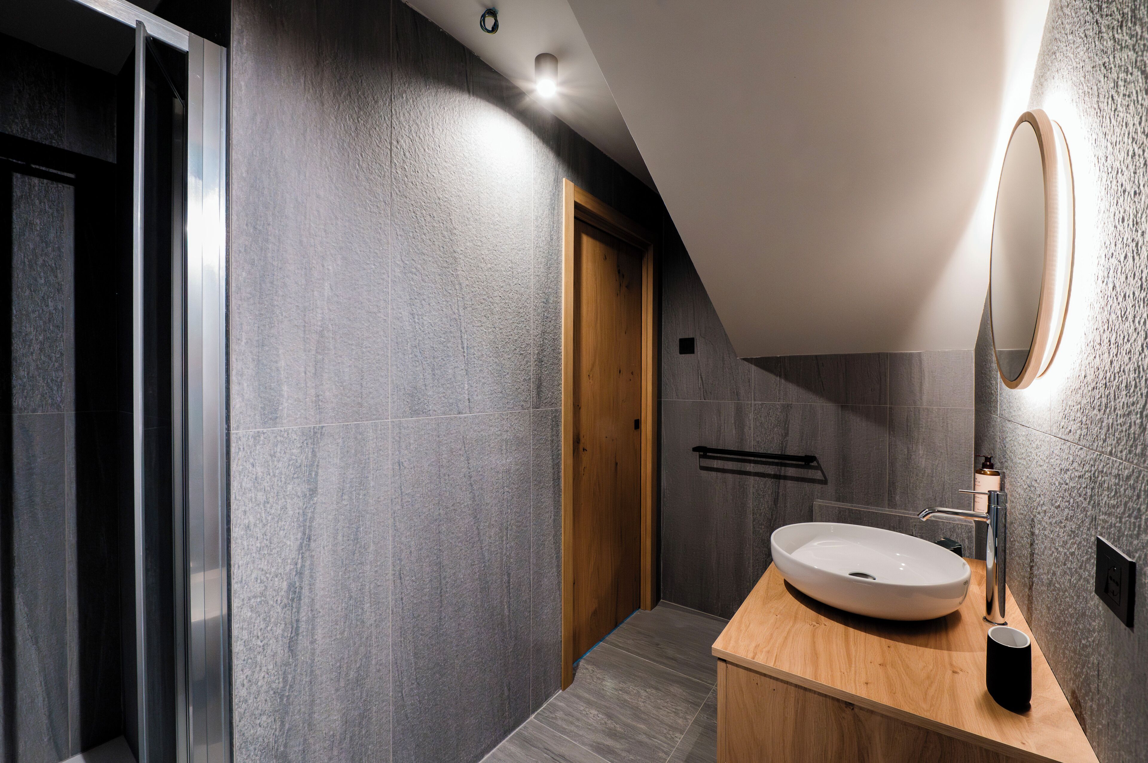A modern bathroom with a wooden door, a large oval sink, and a round mirror.