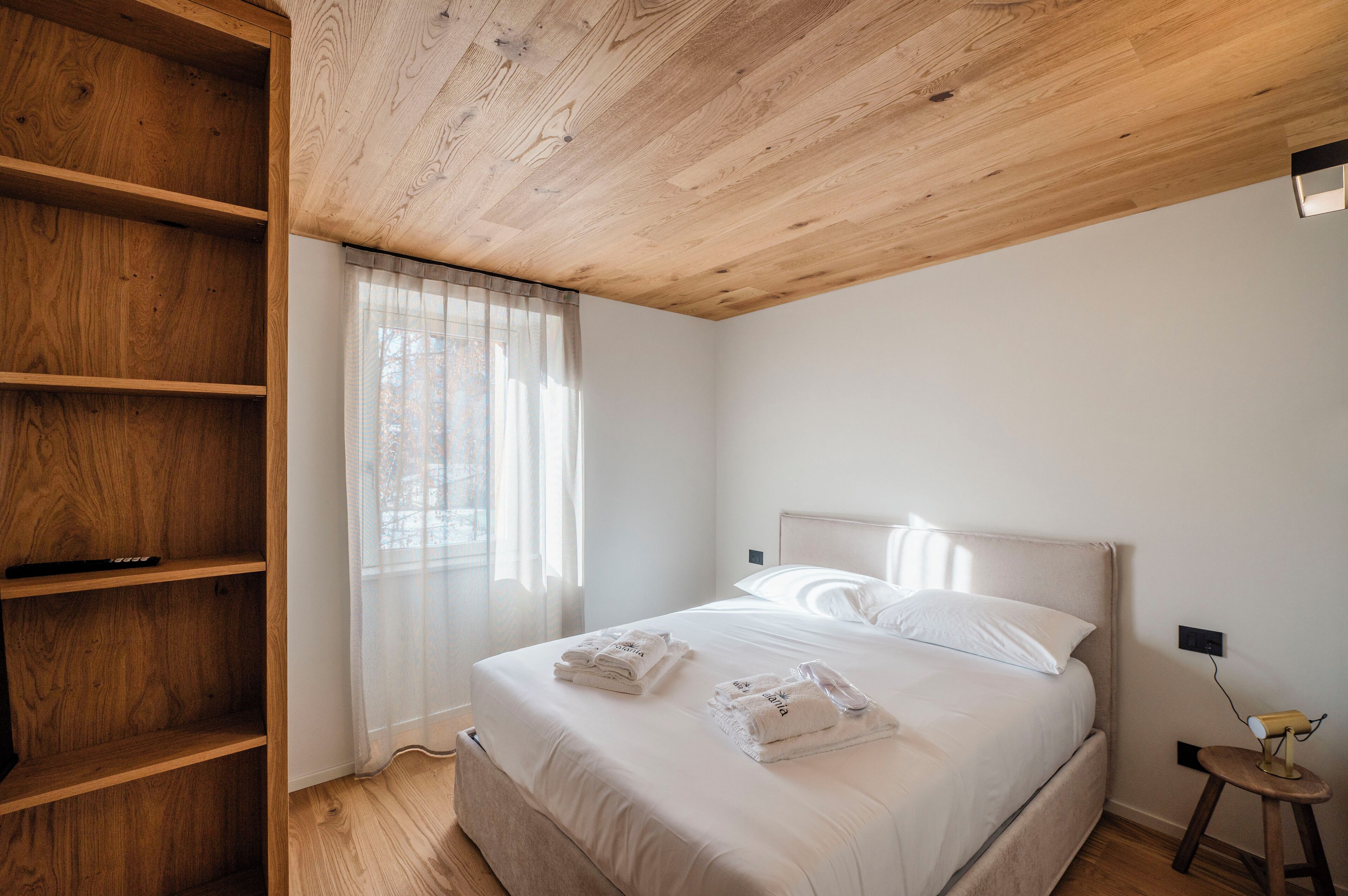 A bedroom with a wooden ceiling, a bed with white linens, a bedside table with a lamp, and a wooden shelf unit.