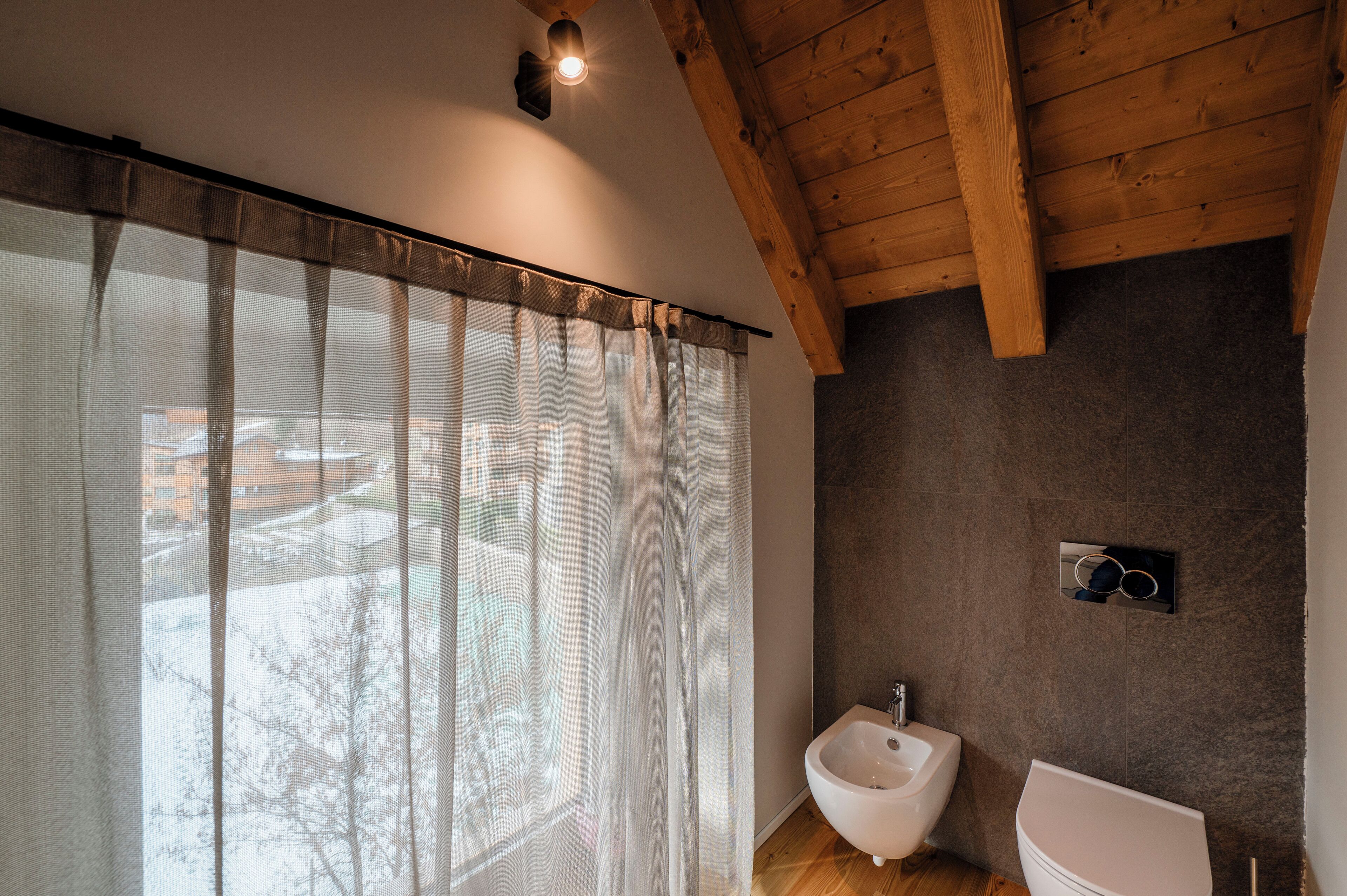 A bathroom with a wooden ceiling, a large window with sheer curtains, a bidet, and a wall-mounted mirror.