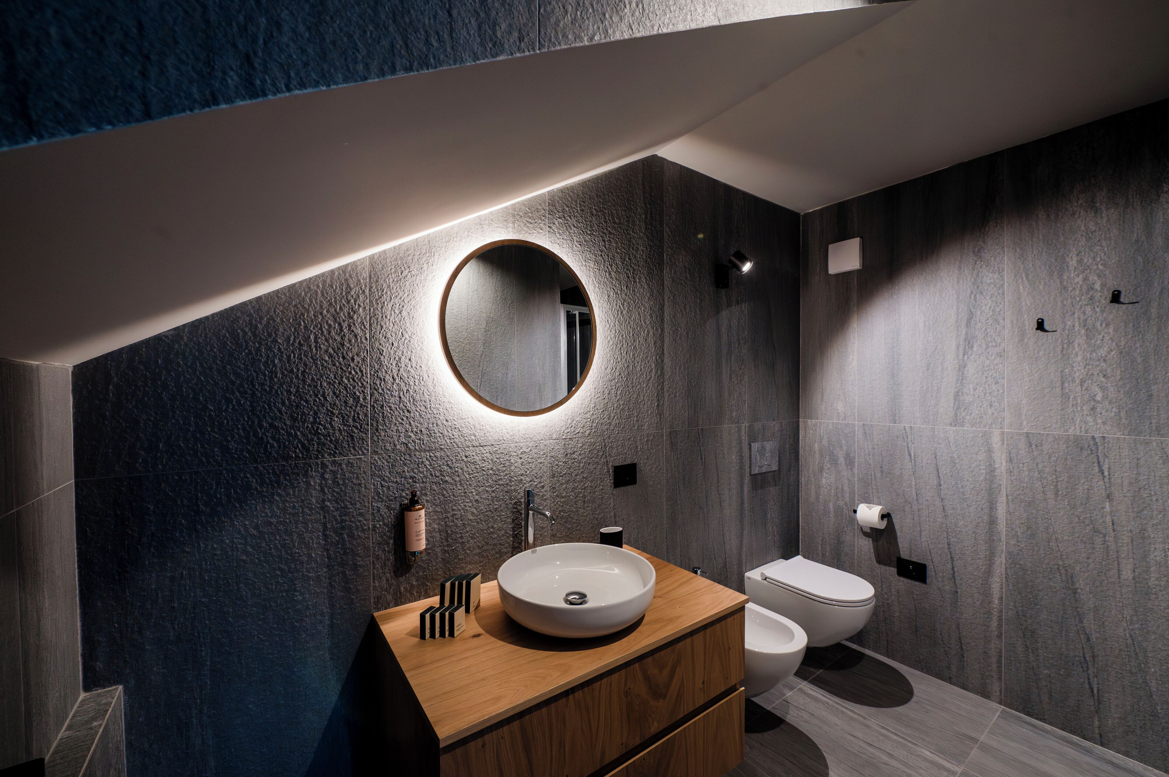 A modern bathroom with a round mirror, wooden vanity, and a toilet.