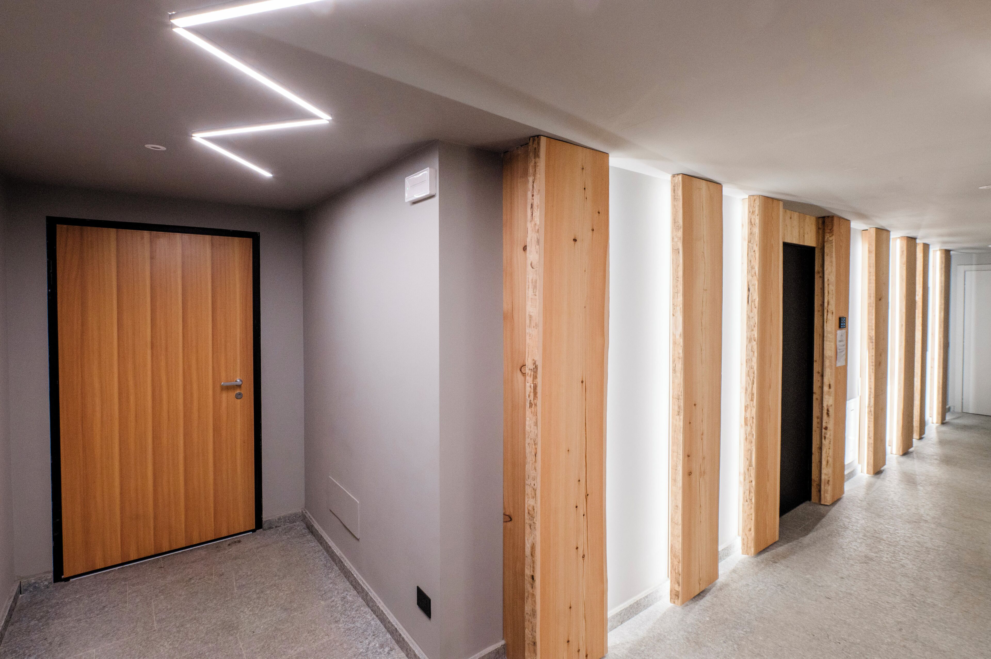 Un couloir aux murs recouverts de lambris en bois, une porte fermée et un plafond doté de luminaires linéaires.