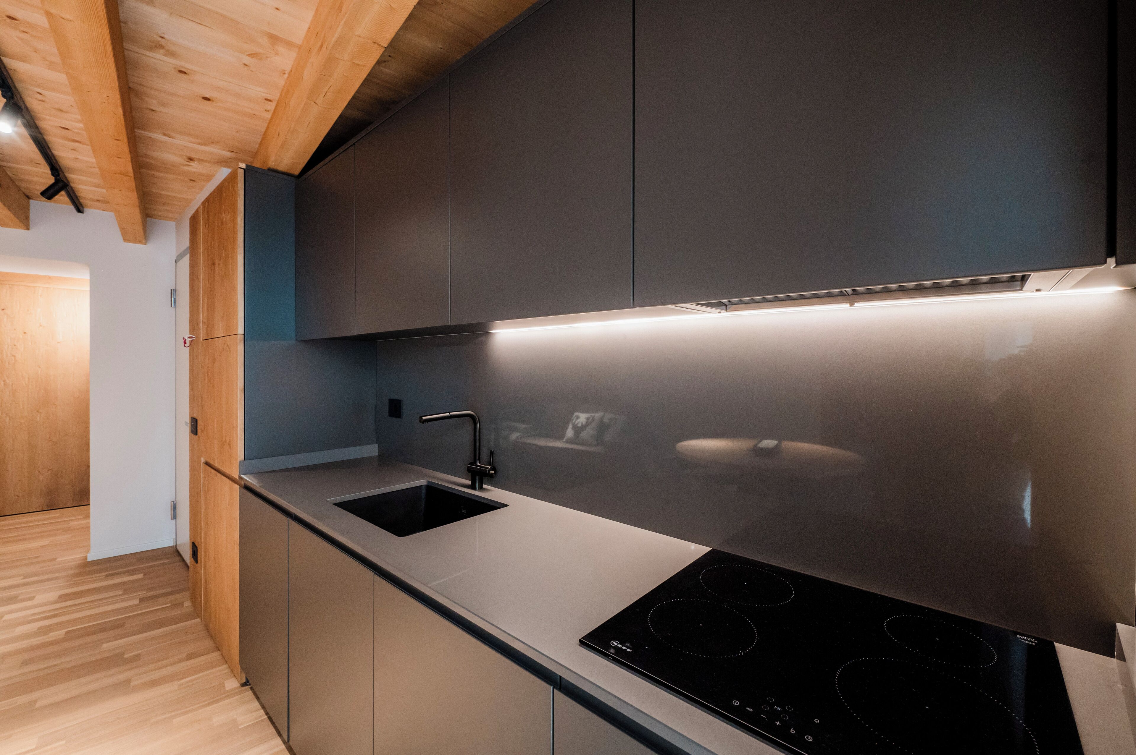 A modern kitchen with a stainless steel sink, a black induction cooktop, and wooden ceiling beams.