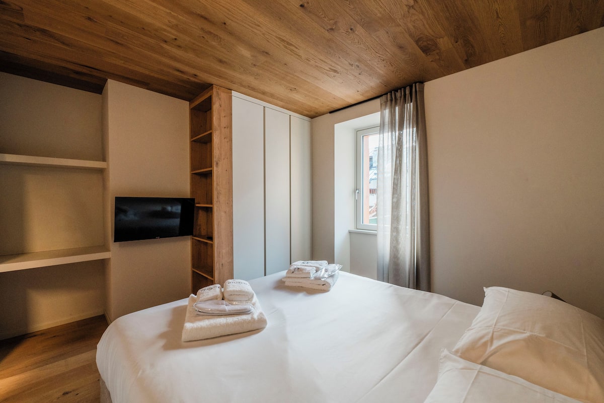Habitación de hotel con techo de madera, cama con ropa blanca, televisor montado en la pared y ventana con cortinas.