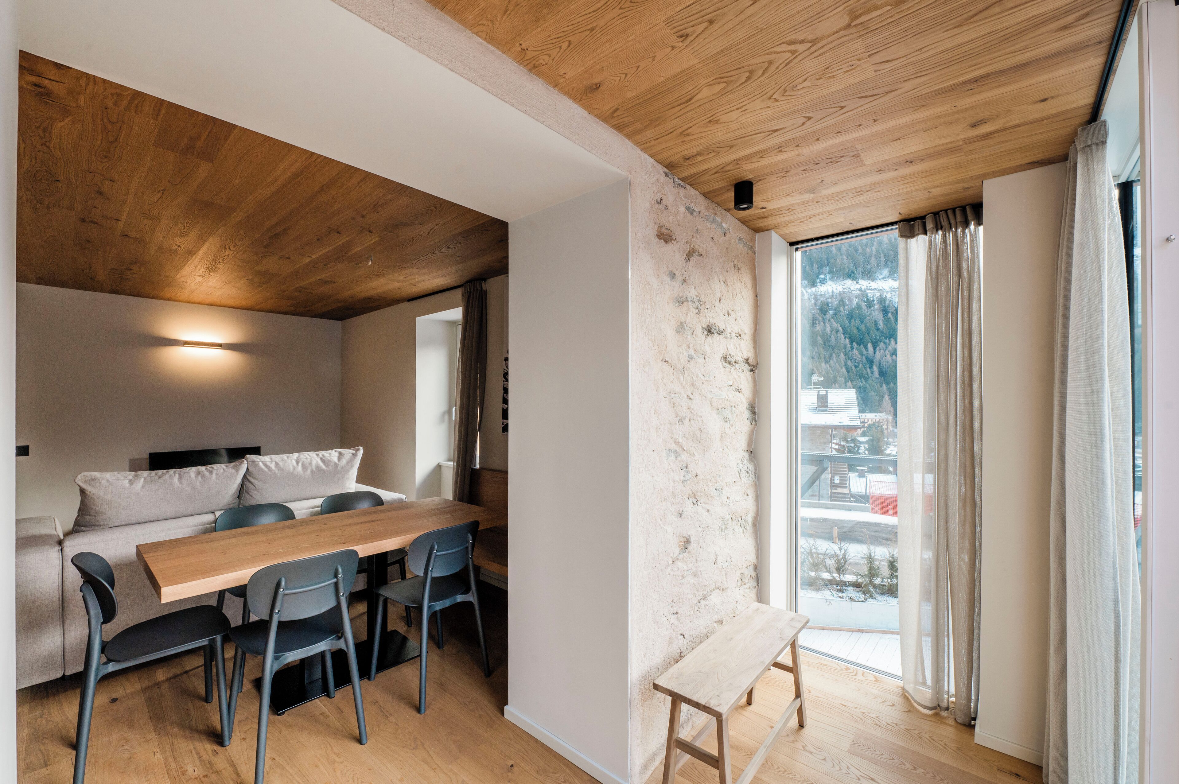 Um interior moderno com teto de madeira, área de jantar com mesa e cadeiras de madeira, um banco e uma janela grande com cortinas.