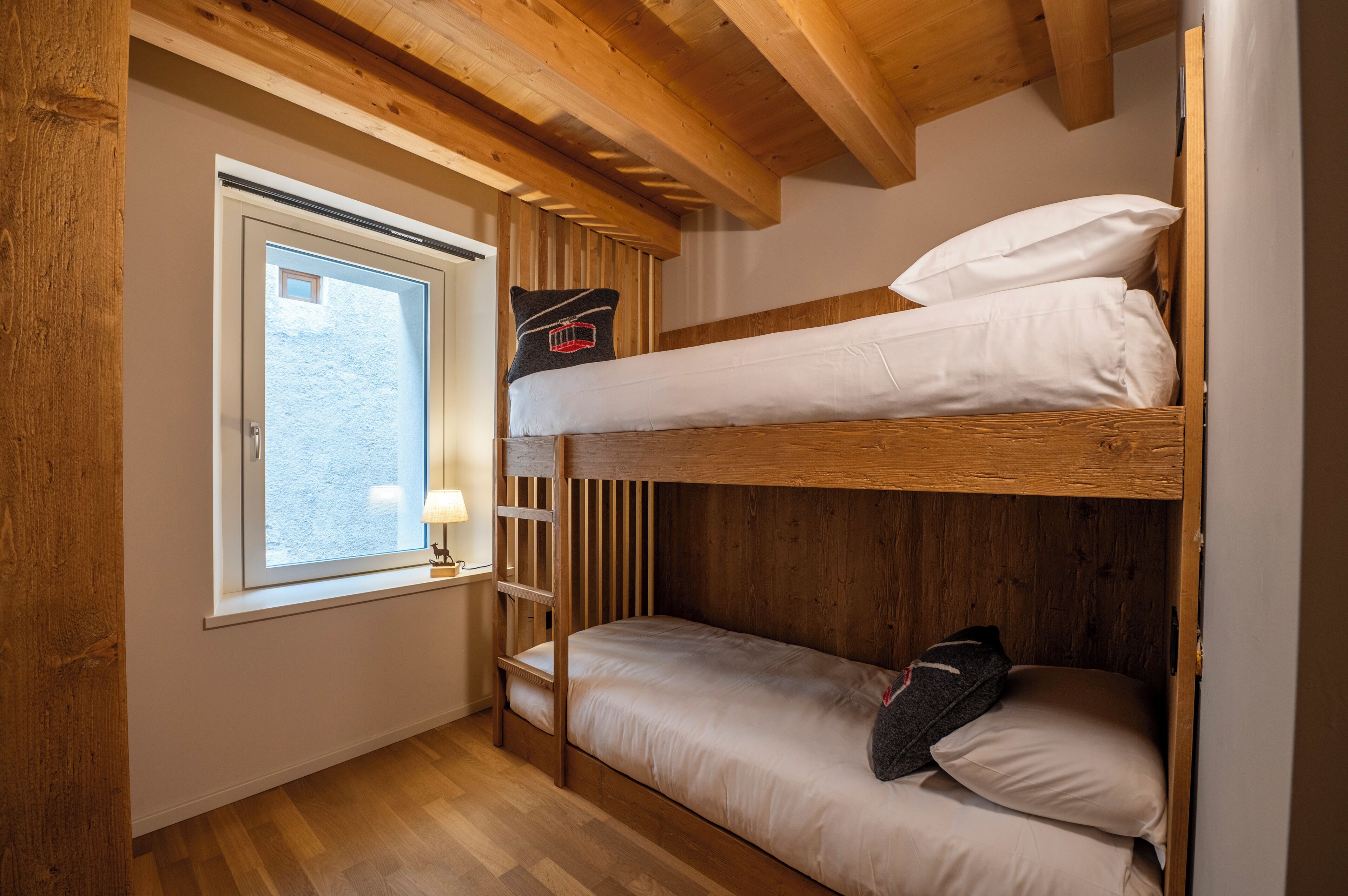 Une chambre avec un lit superposé, une fenêtre, des poutres en bois et une petite table avec une lampe.