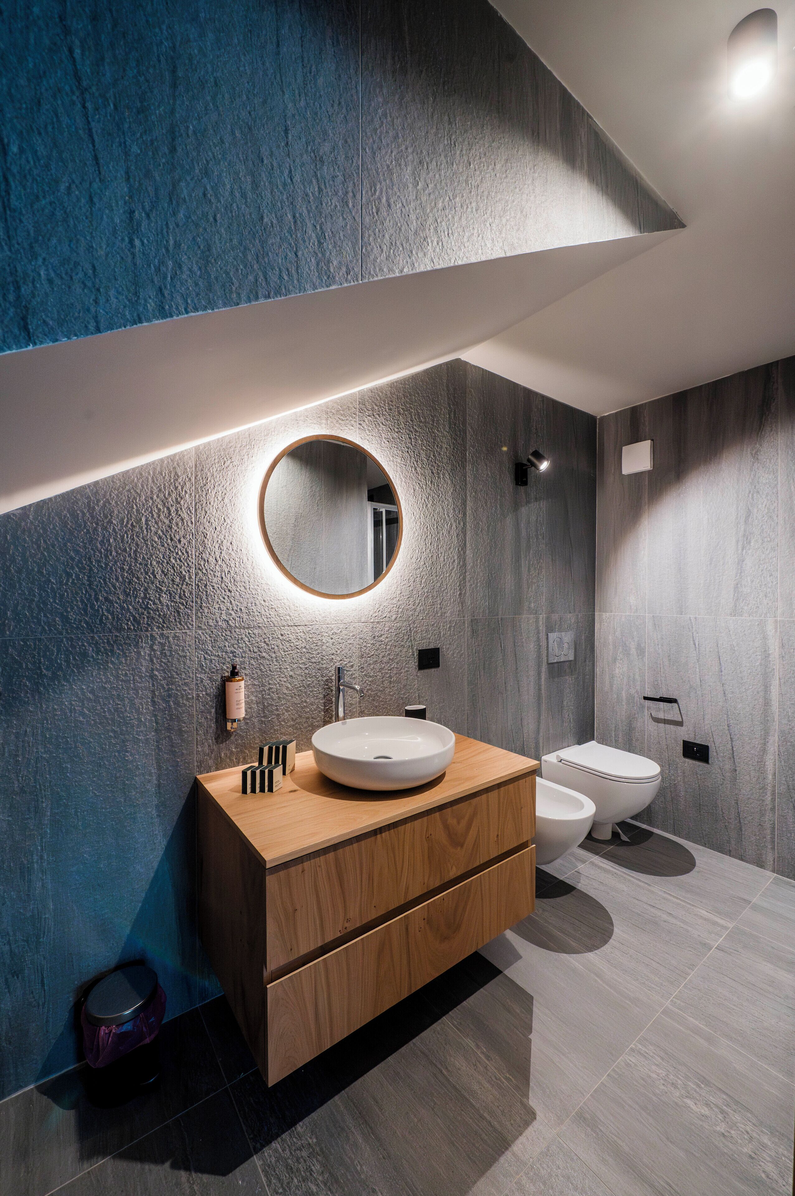 A modern bathroom with a wooden vanity, a round mirror, a white sink, and a toilet.
