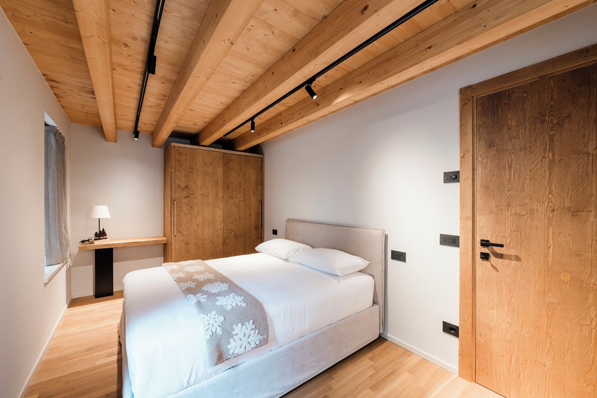 Un dormitorio con una cama, vigas de madera en el techo, un armario y un escritorio con lámpara.