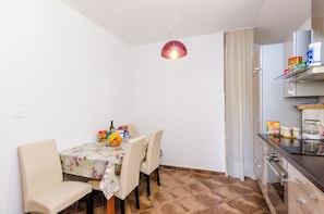 A compact kitchen with a dining area. The kitchen features a modern stove, a coffee maker, and a variety of food items on the counter. The dining area has a table with chairs and a tablecloth. - Ana Apartment (Konavle)