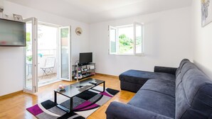 A modern living room with a sofa, a coffee table, and a TV. There is a sliding glass door leading to an outdoor area with a chair and a table. - Ana Apartment (Konavle)