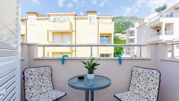 A balcony with two patterned chairs, a small table with a potted plant, and a view of adjacent buildings. - Ana Apartment (Konavle)