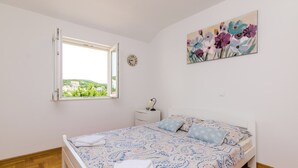 A bedroom with a bed, a window, a bedside table, a clock, and a floral painting on the wall. - Ana Apartment (Konavle)
