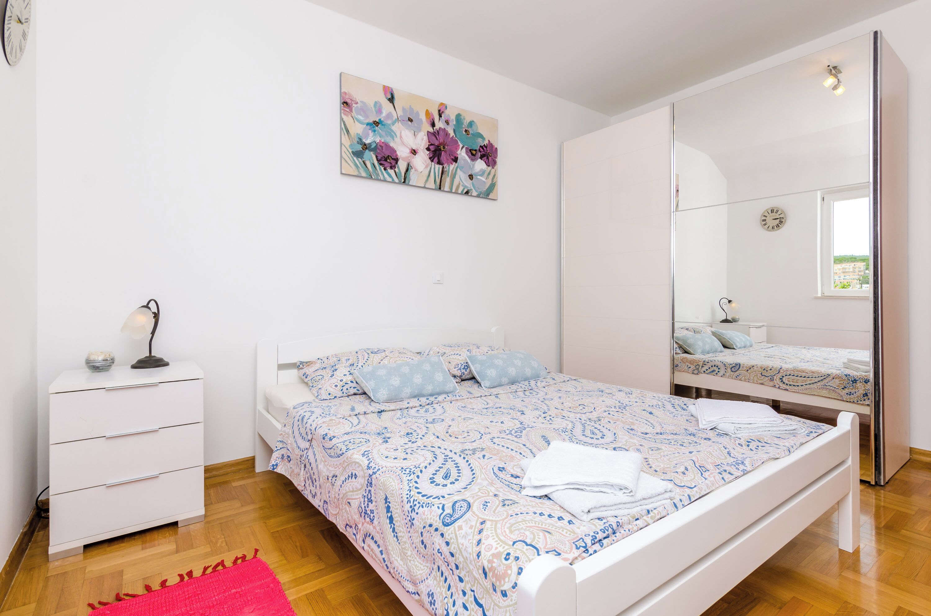 A bedroom with a bed, a nightstand, a lamp, a mirror, and a painting on the wall.