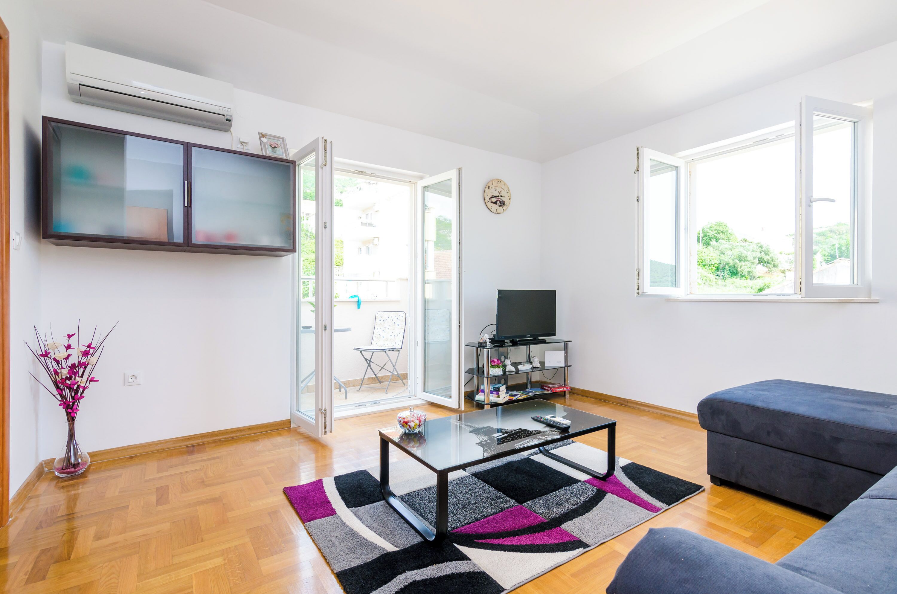 A modern living room with a flat-screen TV, a glass coffee table, a grey sofa, and a large window.