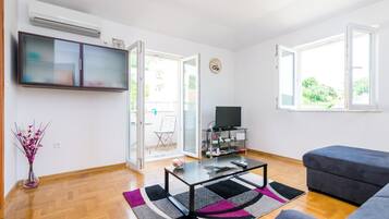 A modern living room with a flat-screen TV, a glass coffee table, a grey sofa, and a large window.