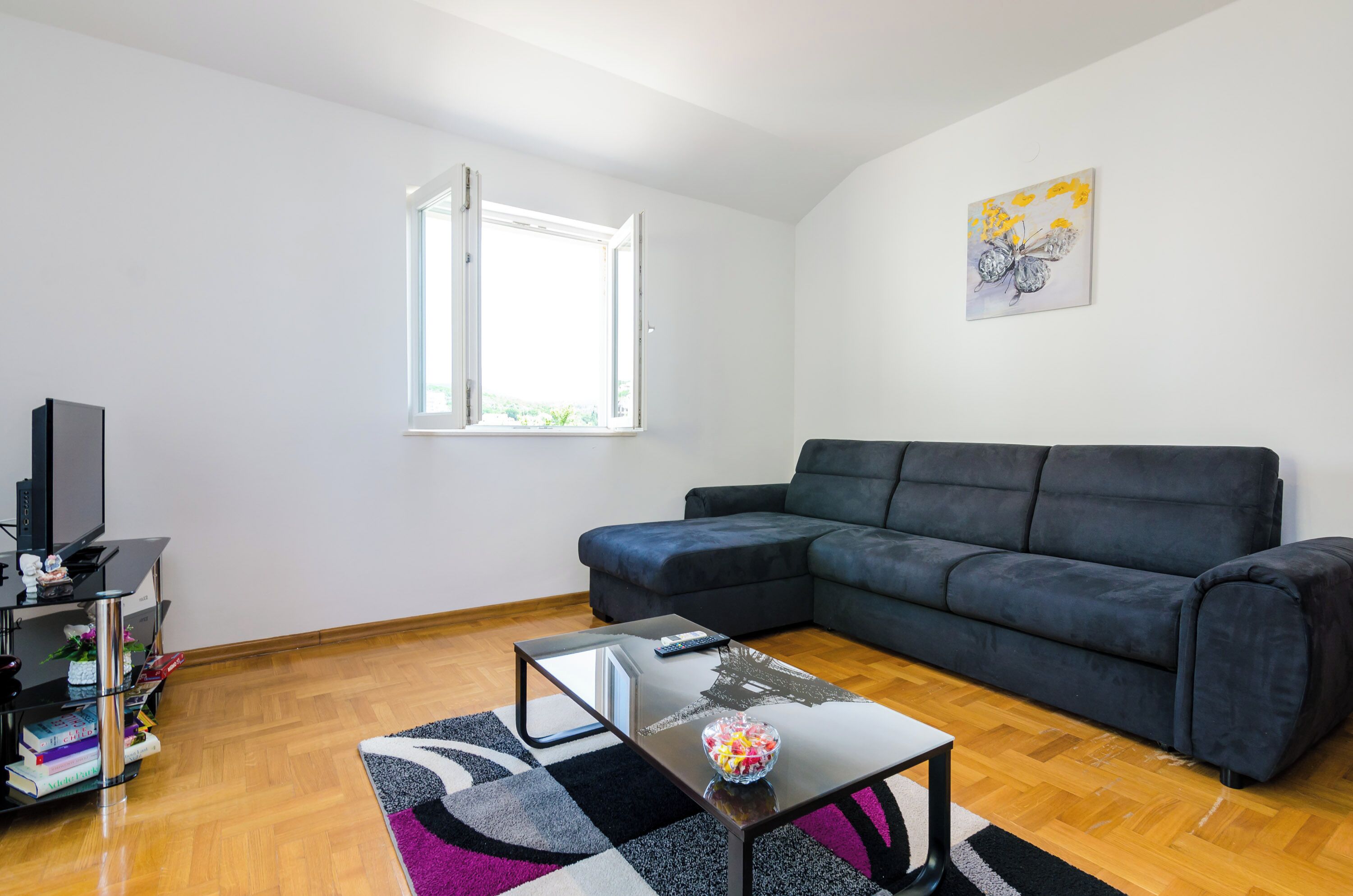 Un salón con sofá de cuero negro, una mesa de centro de vidrio, un mueble para televisión negro y una ventana con vista despejada.