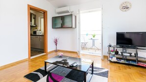 A living room with a glass coffee table, a flat-screen TV, a bookshelf, and a view to a balcony. - Ana Apartment (Konavle)