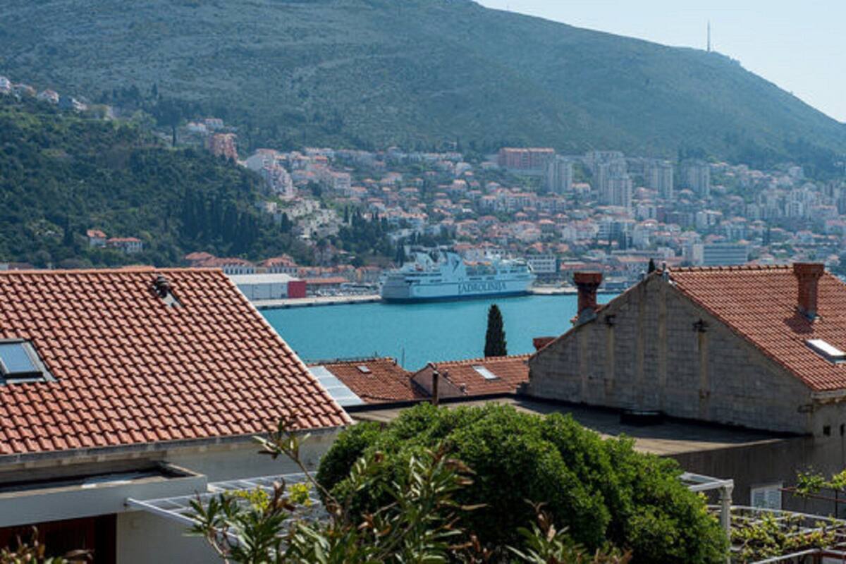 Un paisaje urbano costero con un barco destacado en el puerto, rodeado de edificios residenciales con techos de tejas rojas.