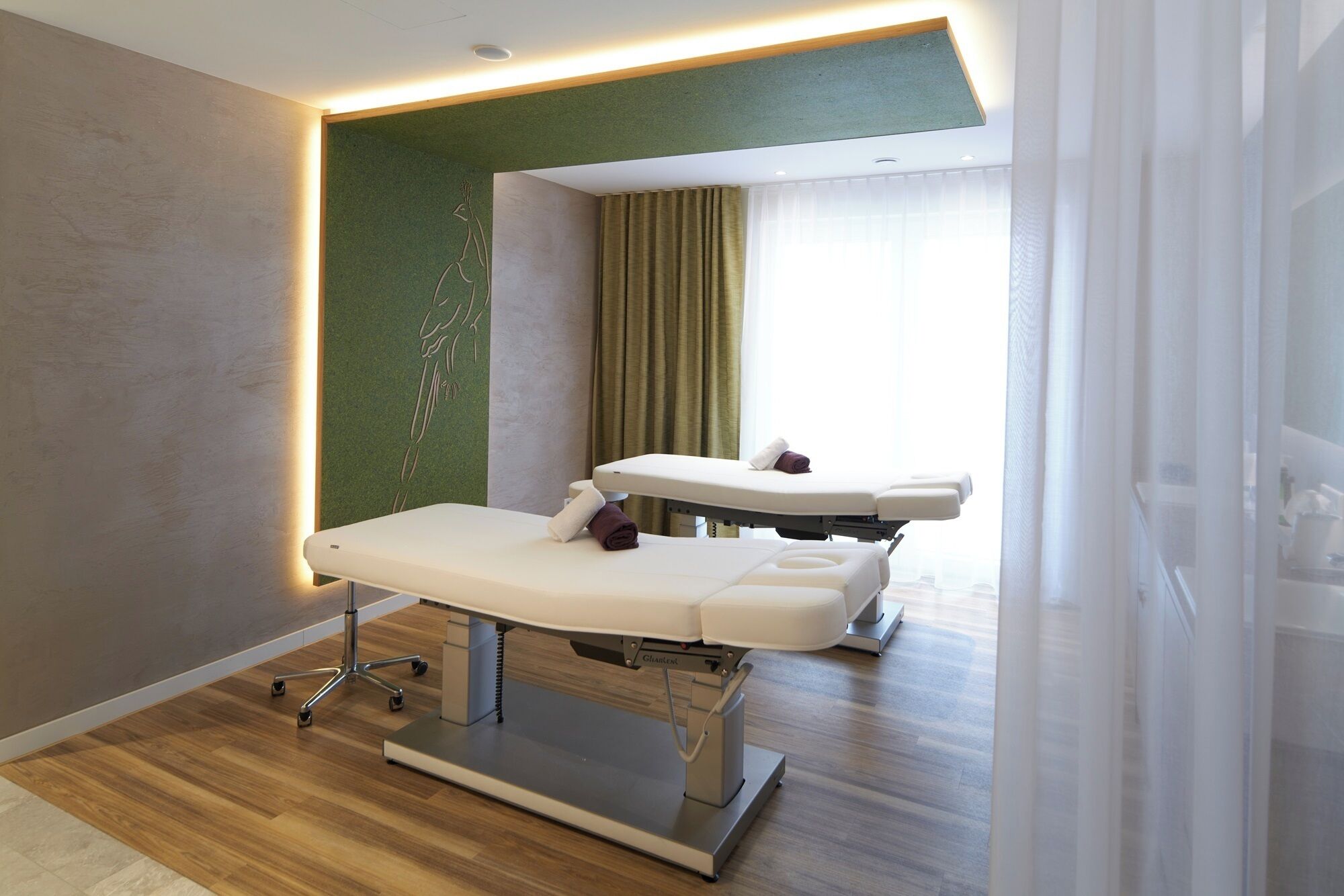 A modern medical examination room with two examination tables, a green accent wall, and a large window with curtains.