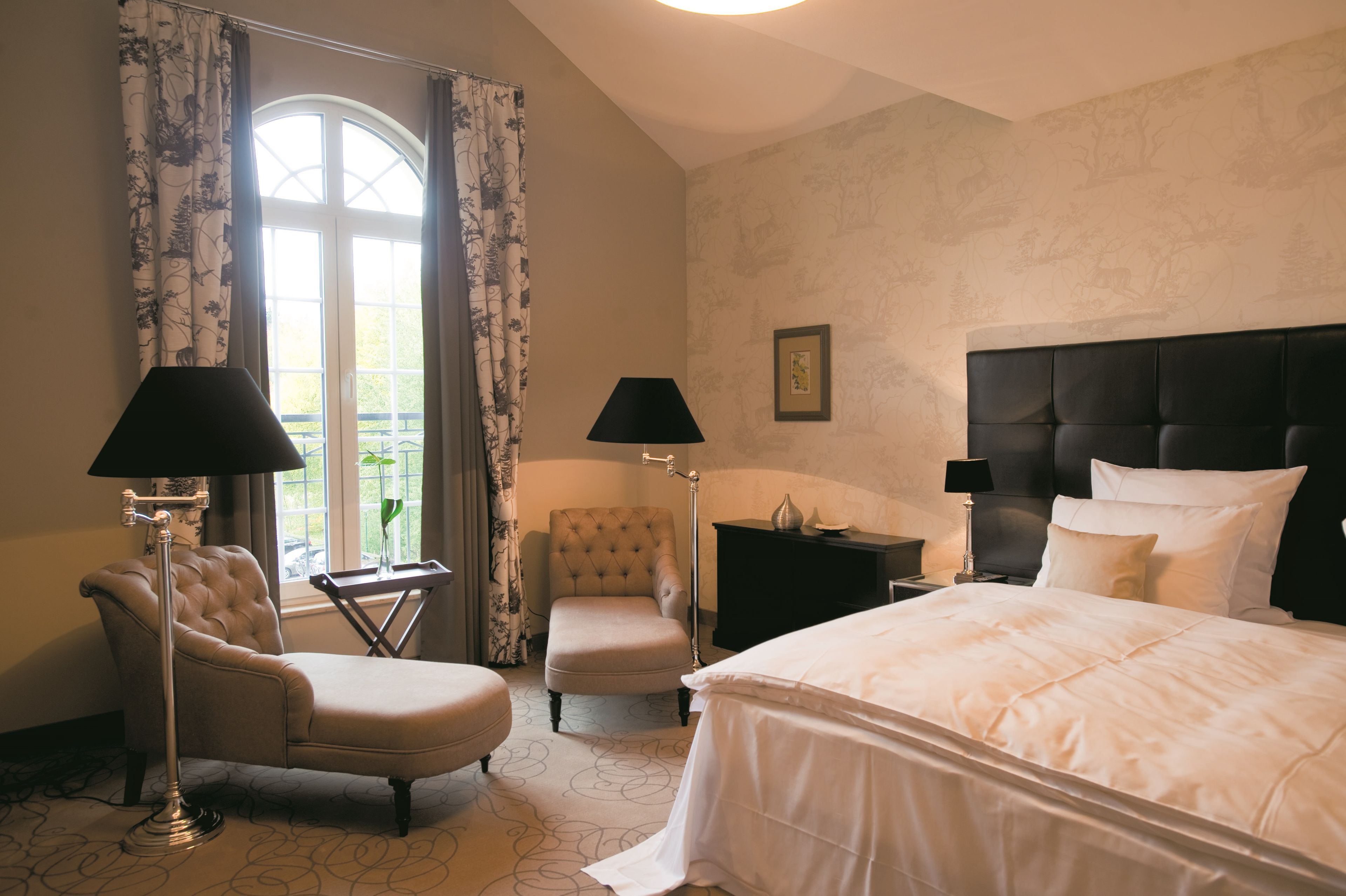 A hotel room with a large bed, two armchairs, a nightstand, and a window with curtains.