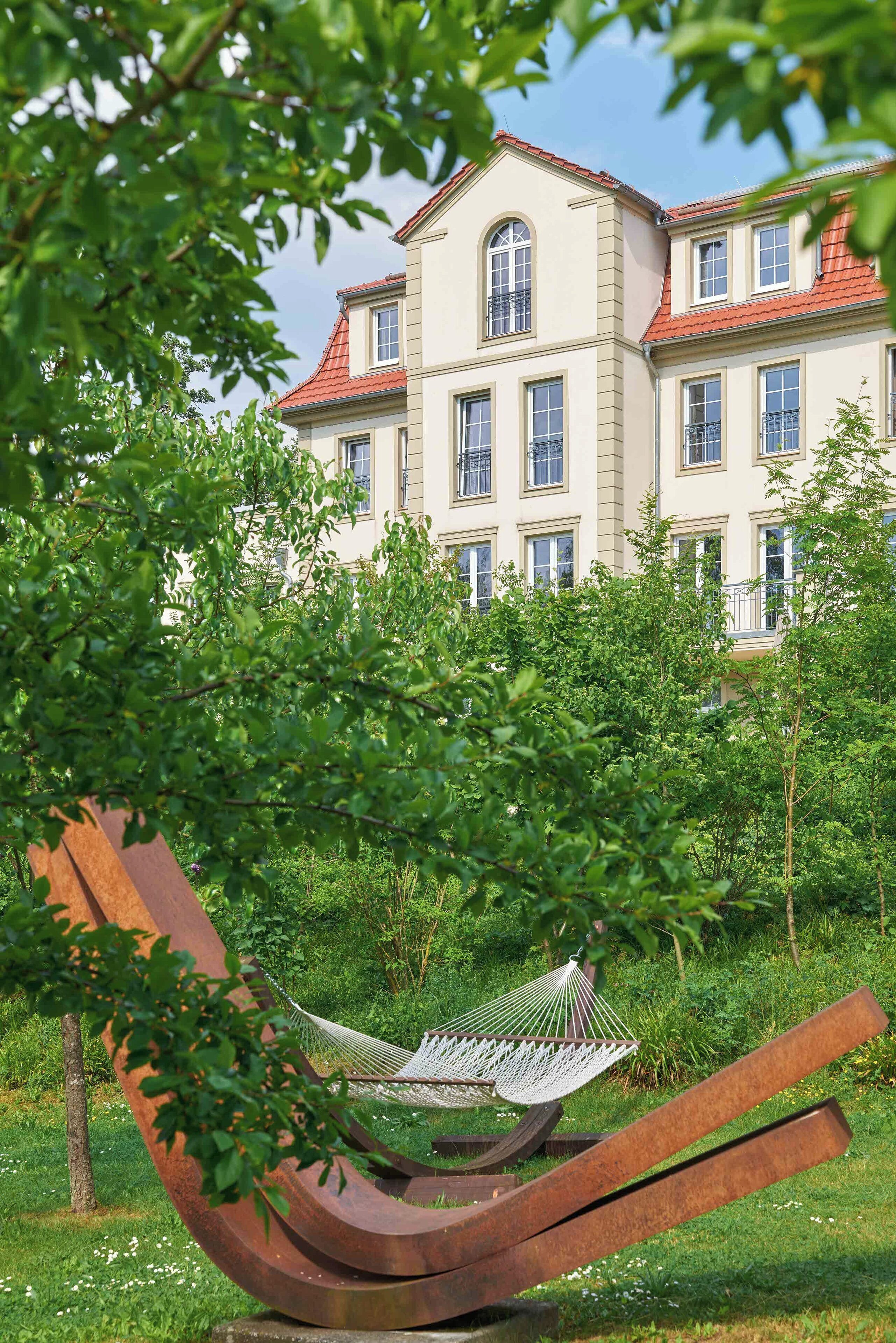 A hammock in a lush garden with a large building in the background.