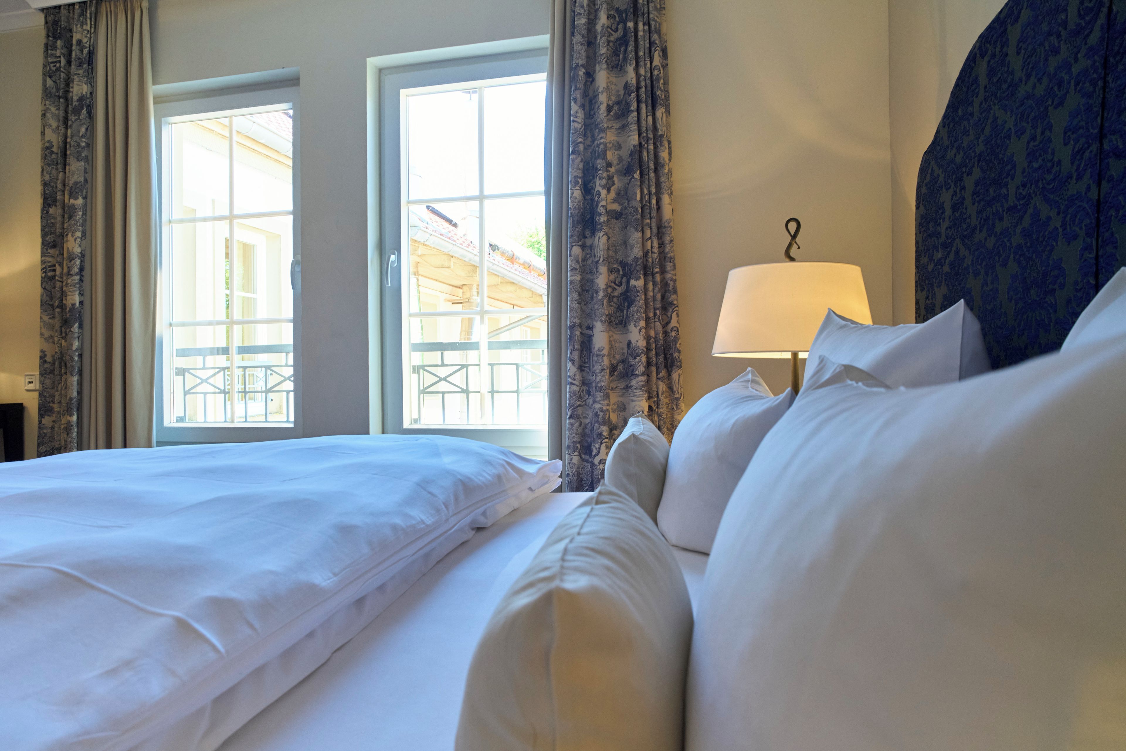 Habitación de hotel con una cama grande, dos ventanas con cortinas, una lámpara y vistas al exterior.