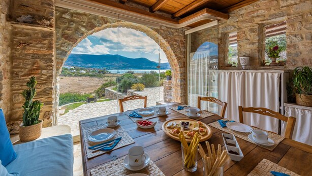 A dining area with a stone wall, a table set for two, and a view of a mountainous landscape. - Blue Greens Villas in Krios, Paros Island (Paros)