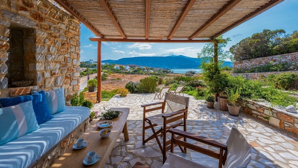A stone-walled room with a wooden bench and a sofa, a table with cups and a bowl, and a view of the sea and mountains. - Blue Greens Villas in Krios, Paros Island (Paros)